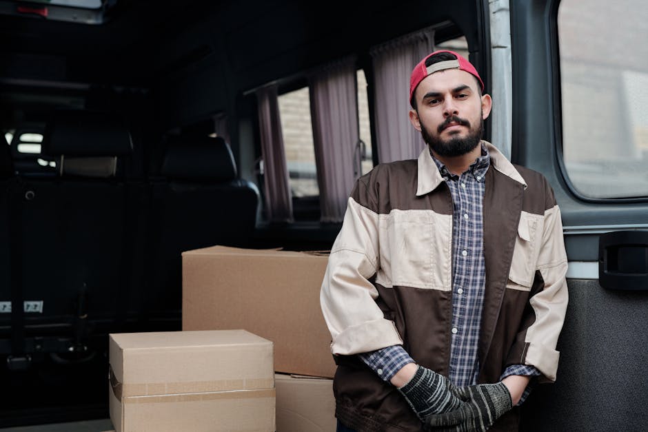 A man with a beard, wearing a red cap, a brown and beige jacket over a checkered shirt, and striped gloves, standing inside a vehicle with a sliding door open, preparing for home relocation. Behind hi