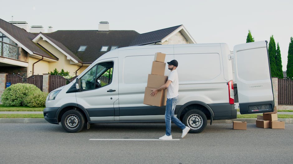 A man with a beard, wearing a red cap, a brown and beige jacket over a checkered shirt, and striped gloves, standing inside a vehicle with a sliding door open, preparing for home relocation. Behind hi
