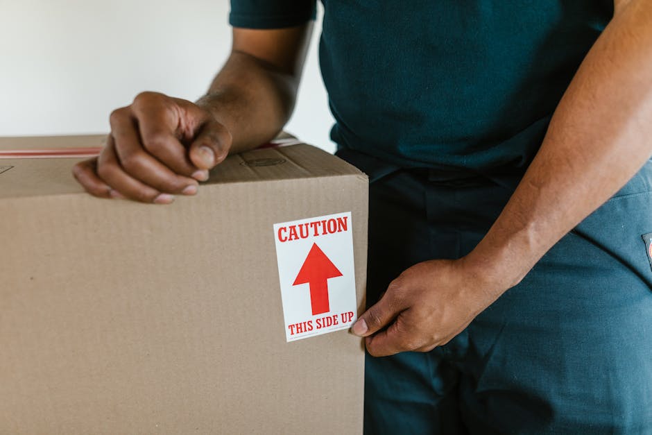 A person wearing a blue work jumpsuit, protective goggles, and pink work shoes stands inside a room with a textured light blue wall and wooden flooring, surrounded by several cardboard boxes used for packing and moving. The individual is holding a pen and appears to be writing or checking a list, suggesting an organized packing or inventory process. One of the boxes has a red label indicating it contains glass items, and they are positioned on or next to larger boxes, some of which have black barcode labels. To the left, a white shelving unit is visible, holding a large potted plant with long green leaves, adding a touch of decor to the space. This scene exemplifies home relocation or packing preparations, with the person focused on ensuring proper documentation before moving, an activity often associated with professional removals services like those offered by Man With a Van Dartford during a house move in the DA1 postcode area.