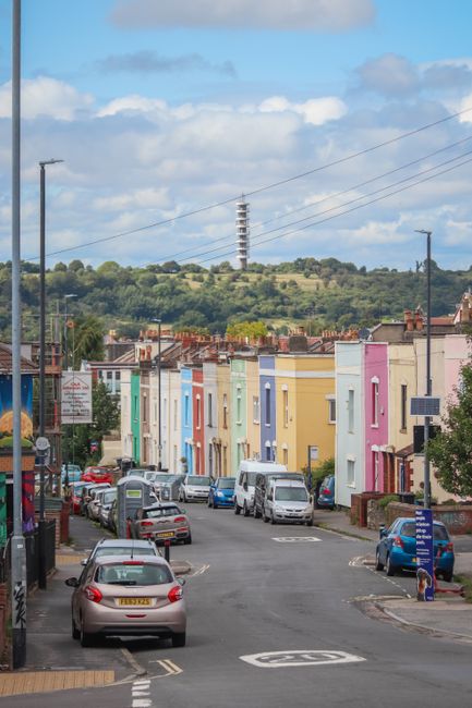 A residential street with a row of colorful terraced houses painted in pastel shades of pink, yellow, blue, and green, positioned closely along the pavement. Several parked cars, including a silver hatchback in the foreground and other vehicles further down the street, occupy designated parking bays and partly extend onto the road. The street's surface is asphalt with visible road markings, and there are utility poles with overhead wiring running parallel to the houses. In the background, a hill with lush greenery and trees features a tall electrical pylon, against partly cloudy skies. The scene captures a typical urban environment suitable for home relocation or furniture transport, with the surrounding environment indicating a quiet residential area that [COMPANY_NAME], such as Man With a Van Dartford, might service for removals or moving logistics. The image is well lit with natural daylight, emphasizing the clarity of the parked vehicles, the vibrant house facades, and the overall setting for loading or unloading activities involved in packing and moving tasks.
