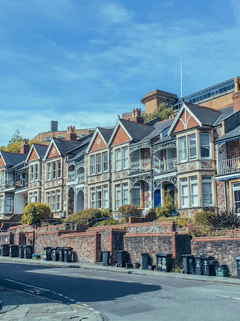 A row of Victorian-style terraced houses on a residential street with brick facades, bay windows, and decorative ironwork on small balconies. The houses are situated on a raised pavement with a low brick retaining wall, behind which are neatly maintained shrubs and small trees. In the foreground, black wheeled rubbish bins are lined up along the edge of the street, which has a marked parking area and some parked vehicles further down. Above, the sky is clear with a few wispy clouds, and a tall building or tower is visible behind the row of houses. The scene illustrates a typical urban setting potentially relevant to house removals, with a focus on the external environment where moving services like loading or parking near the property may be involved. This visual context supports understanding of house access and logistics in Dartford, as part of a home relocation process managed by Man With a Van Dartford.